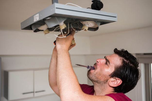 Free photo man carrying tester in mouth while repairing ceiling focus light at home