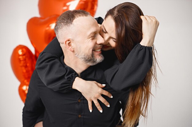Man carrying woman piggyback while posing in studio near bunch of heartshaped red ballons