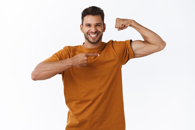 man in casual brown t-shirt, raising hand, tighten muscle and pointing at biceps with satisfied, proud expression