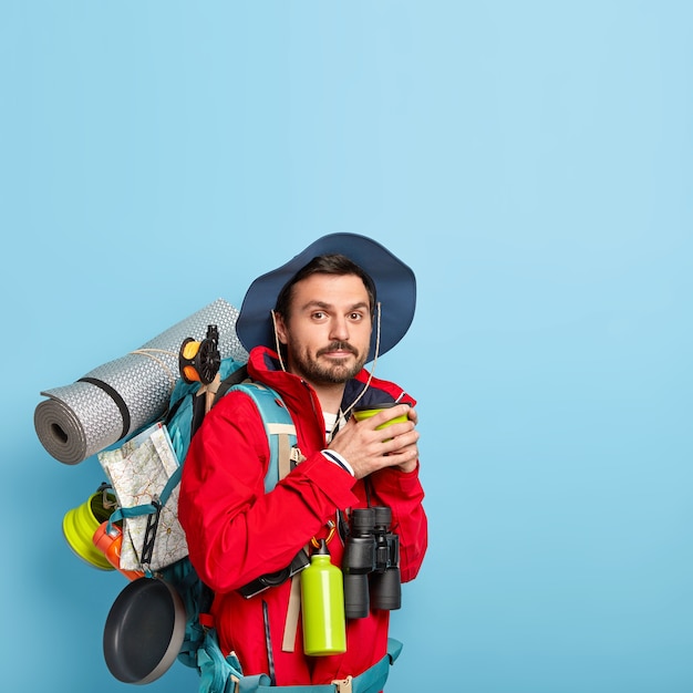 Uomo in abbigliamento casual, beve caffè, trascorre il tempo libero, porta karemat, tiene un binocolo, isolato sul muro blu, copia lo spazio sopra cerca avventure