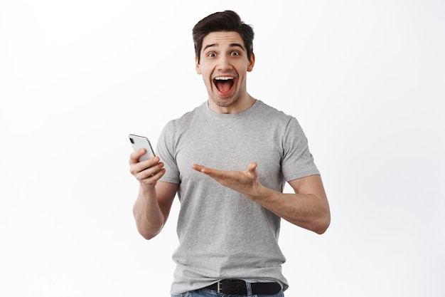 携帯​電話​で​素晴らしい​ニュース​を​祝う​男​は​、​興奮して​見て​、​白い​背景​の​上に​立って​、​うれしそう​な​驚き​の​顔​で​スマート​フォン​の​画面​を​指しています