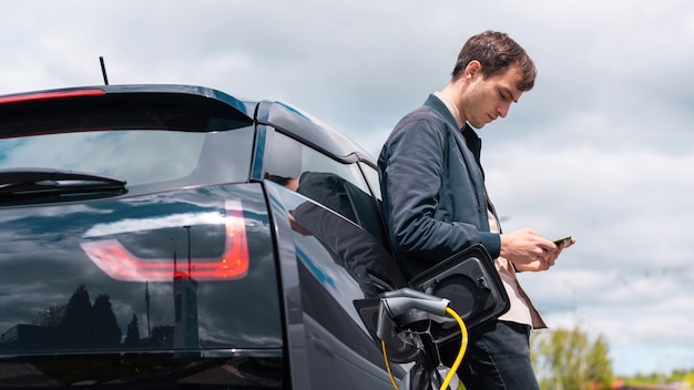 Человек заряжает свой электромобиль на зарядной станции и использует смартфон