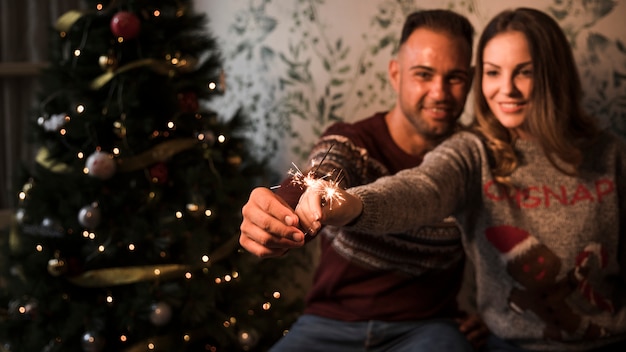 Man and cheerful woman with flaming Bengal lights near Christmas tree