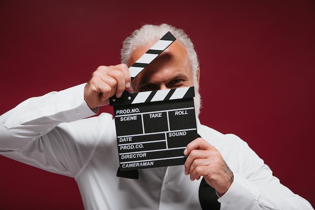 Man in classic style shirt holds clapper White haired funny guy with beard in light clothes posing on burgundy background