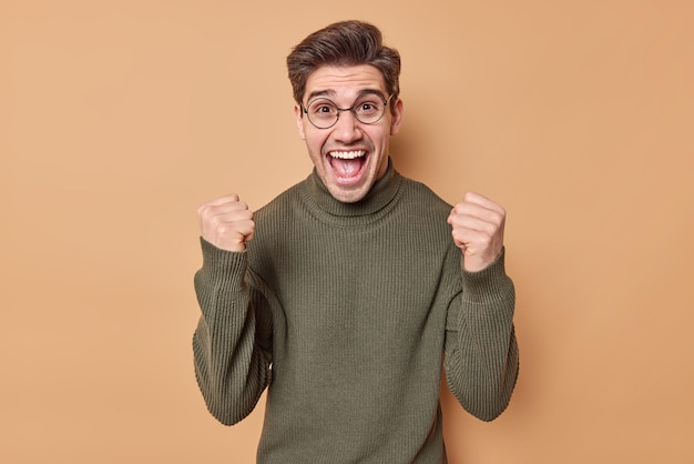 man clenches fists triumps from successful deal or great news being joyful and thrilled wears casual jumper and spectacles poses agaist beige catches dream