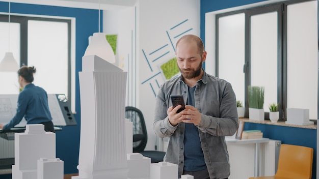 Man contractor using mobile phone to design building model for construction plan and layout. Architect working blueprints plan and maquette to create industrial development on smartphone.