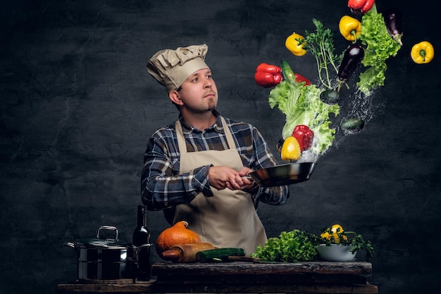 Мужчина-повар держит сковороду с овощами, летящими в воздухе.
