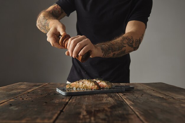 Man cooks healthy meal on rustic wooden table, peppers two raw pieces of salmon in white wine sauce with spices and herbs presented on marble deck prepared for grill