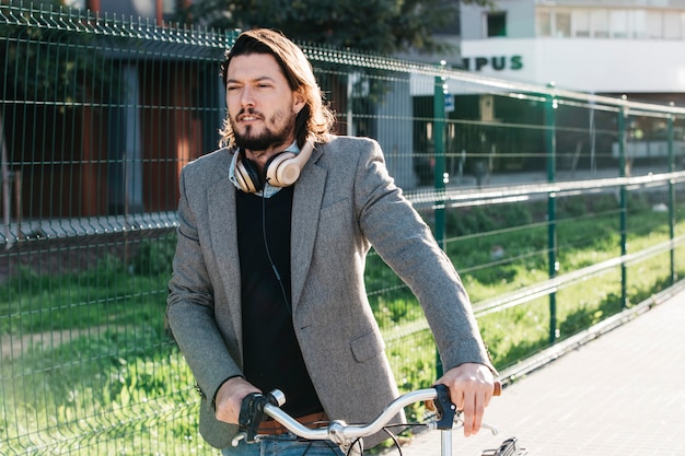 A man in court with headphone around his neck walking with bicycle at outdoors