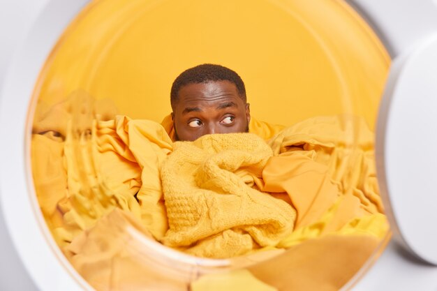  man covered with laundry looks away surprisingly prepares for washing poses inside of washer at home laundromat 