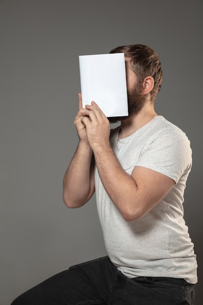 man covering face with book while reading on grey wall