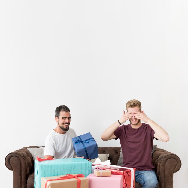 Man covering his eyes with hands sitting on sofa with his friend holding gift box