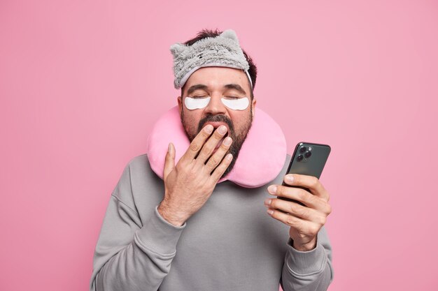  man covers mouth with hand wants to have rest scrolls social networks via smartphone applies patches to reduce puffiness under eyes wears sleepmask travel pillow around neck.