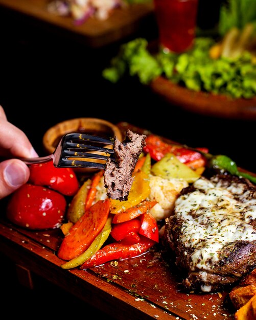 Man cut a piece of beef steak topped with melted cheese served with roasted vegetables