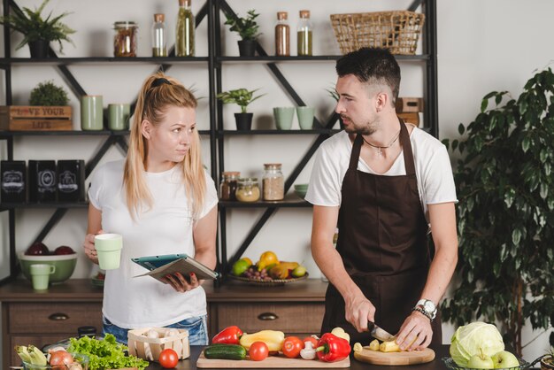 Man cutting vegetables with knife looking at woman holding digital tablet