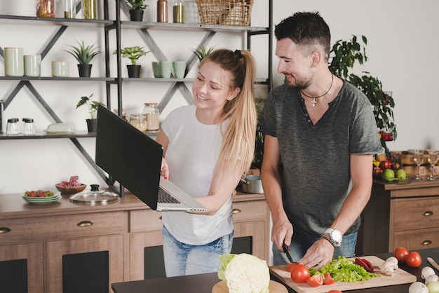 Человек резки овощей с ножом, глядя на женщину, держащей ноутбук на кухне