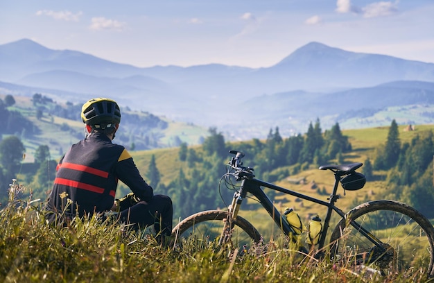 Man cyclist sitting on grass and looking at mountains