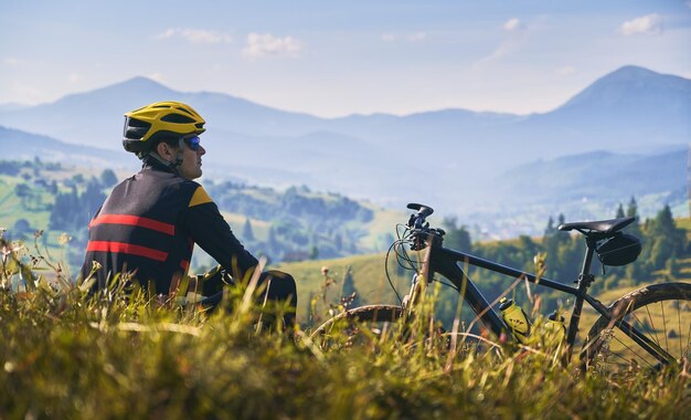 Мужчина-велосипедист сидит на траве и смотрит на горы