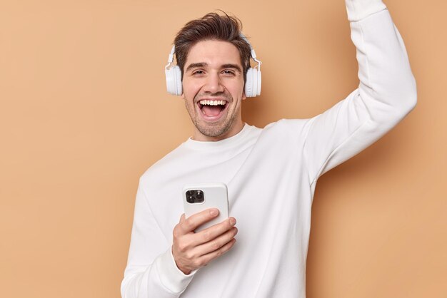 man dances and laughs happily enjoys favorite playlist listens music via headphones holds mobile phone feels joyful isolated on beige boosts mood with favorite song