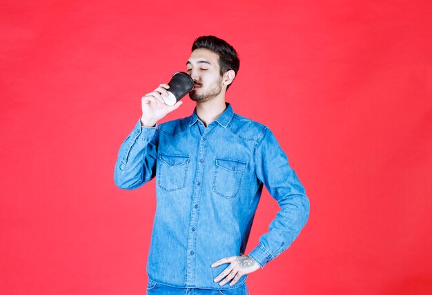 Мужчина в джинсовой рубашке держит черную одноразовую чашку с напитком и пьет ее.
