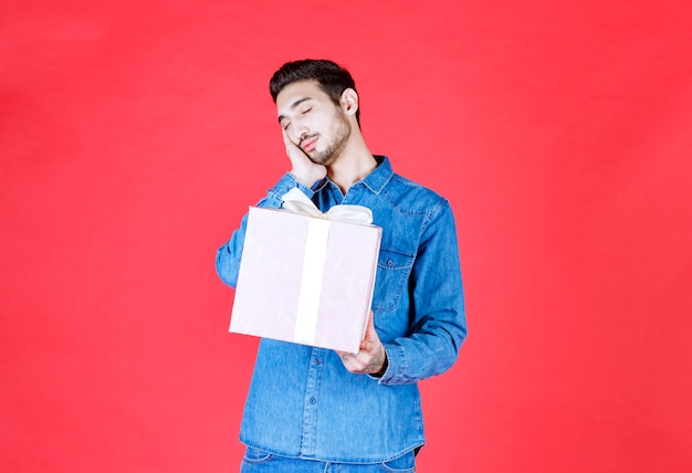 Мужчина в джинсовой рубашке держит фиолетовую подарочную коробку, перевязанную белой лентой, и выглядит сонным и усталым.