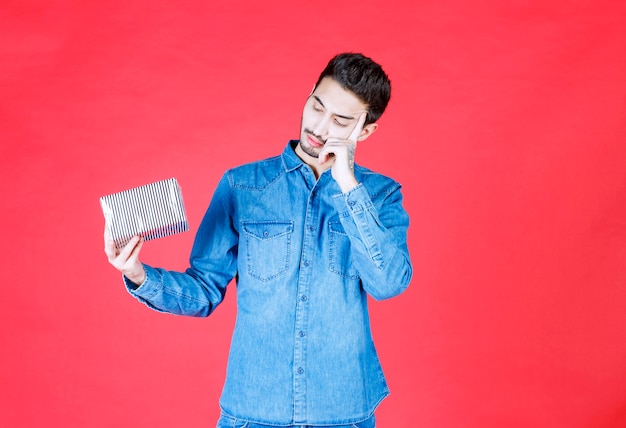 Uomo in camicia di jeans che tiene in mano una confezione regalo d'argento e sembra confuso e inaspettato.