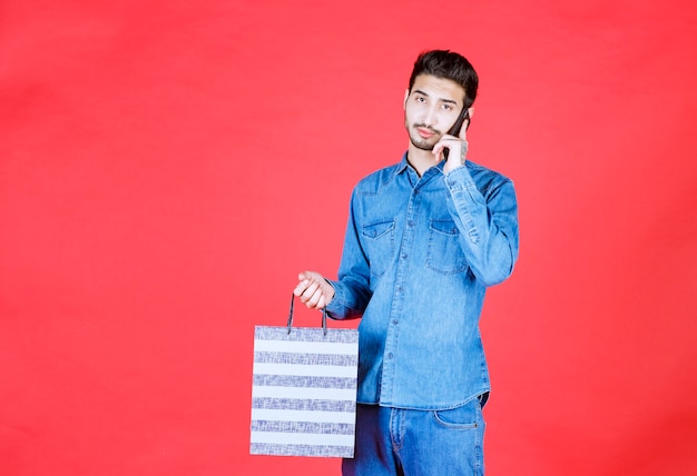 Man in denim shirt holding a striped shopping box and talking to the phone.