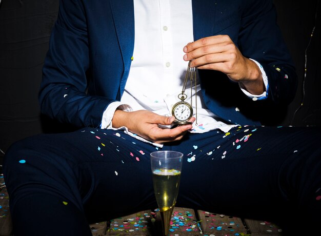 Man in dinner jacket holding pocket watch near glass between confetti 