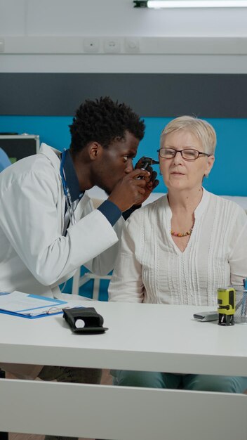 机に座っている高齢患者の耳の検査に耳鏡を使用している男性医師。病気の老婆のための相談を行う医療キャビネットの専門ツールを持つ若い耳科医