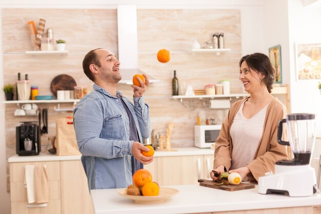 Man doing tricks with oranges for wife in kitchen while preparing healthy smoothie. Healthy carefree and cheerful lifestyle, eating diet and preparing breakfast in cozy sunny morning