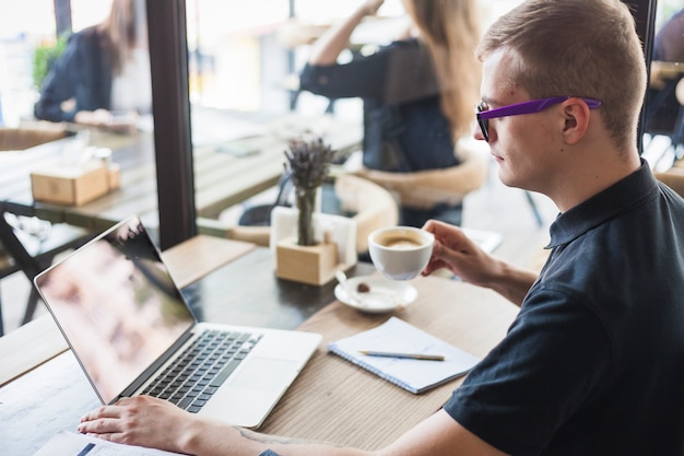 Free photo man drinking coffee at wooden table with laptop