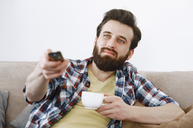 Man drinks coffee. Guy watching tv on couch. TV remote control in hands.