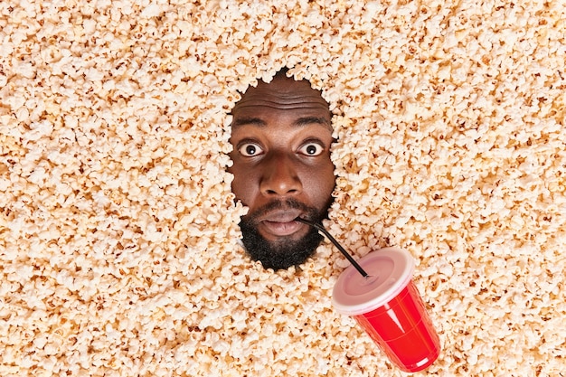 Free photo man drowned in popcorn drinks soda watches film in cinema has surprised expression frightened beacause of horror scene in movie looks excitedly