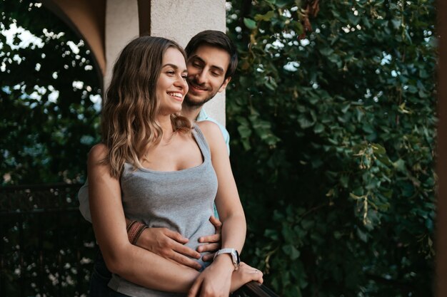 Man embracing wife at balcony. Relaxed couple enjoy day and good news. Happy young family