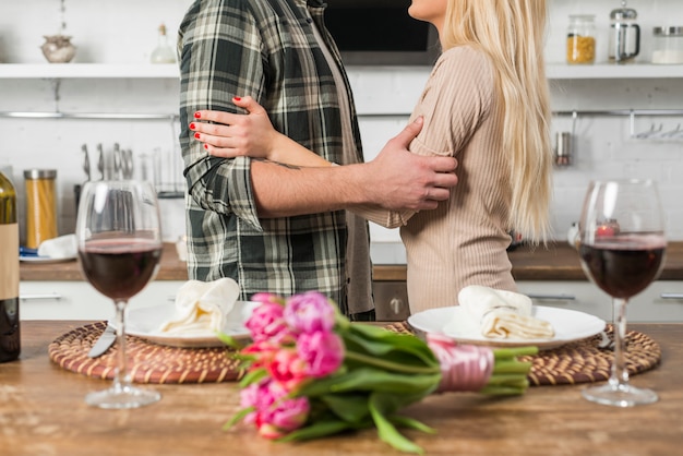 花とワインのグラスを持つテーブルの近くの女性と抱きしめる男