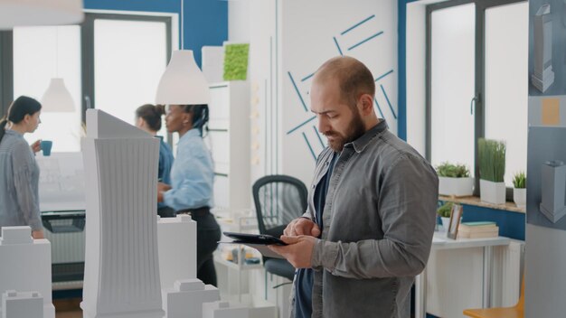 Man engineer designing architectural plan on tablet with building model and maquette. Architect working on urban project to develop property construction layout. Industrial architecture