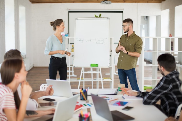 Man in eyeglasses and shirt and smiling woman in blouse standing near board while dreamily presenting new project to colleagues Group of young people working together in modern white office