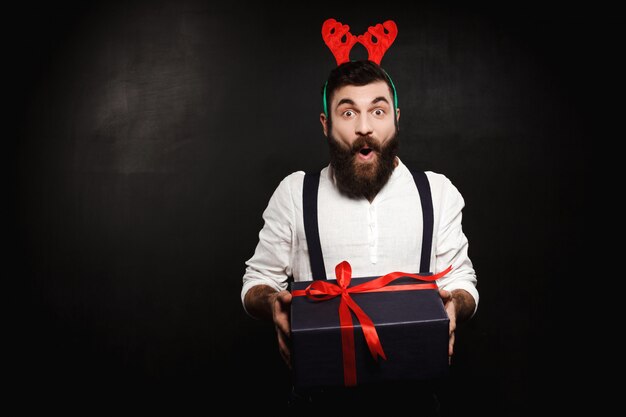 Man in fake deer horns holding christmas gift box over black.