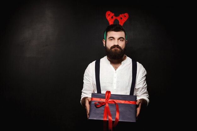 Man in fake deer horns holding christmas gift box over black.