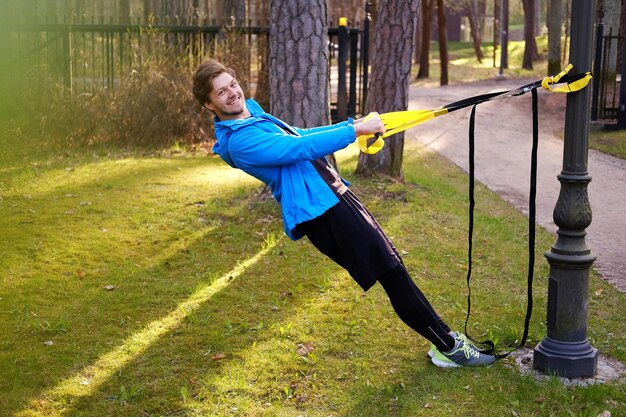 Man fitness model in a blue sport jacket doing workouts with fitness trx strips in a park.