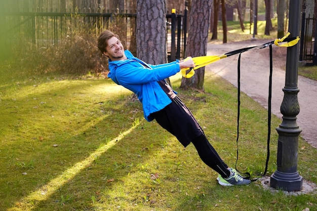 Man fitness model in a blue sport jacket doing workouts with fitness trx strips in a park.