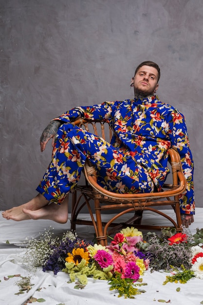 Мужчина в цветочном платье сидит на стуле с разными цветами на полу