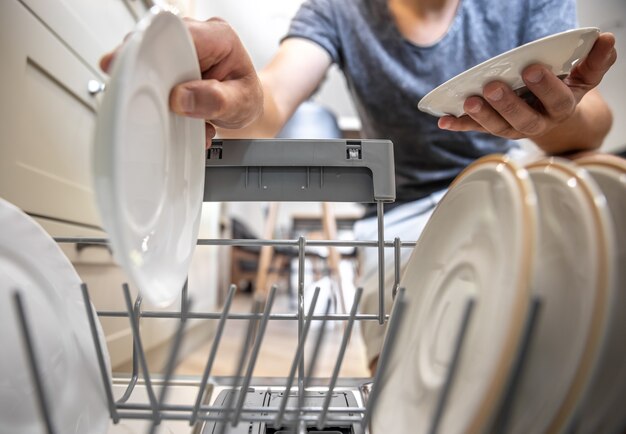 Мужчина перед открытой посудомоечной машиной достает чистую посуду после мытья.