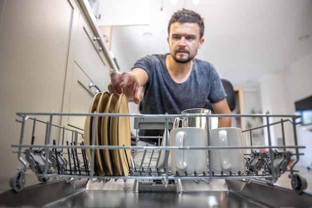 Мужчина перед открытой посудомоечной машиной достает или ставит посуду.