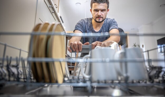 A man in front of an open dishwasher takes out or puts down dishes.