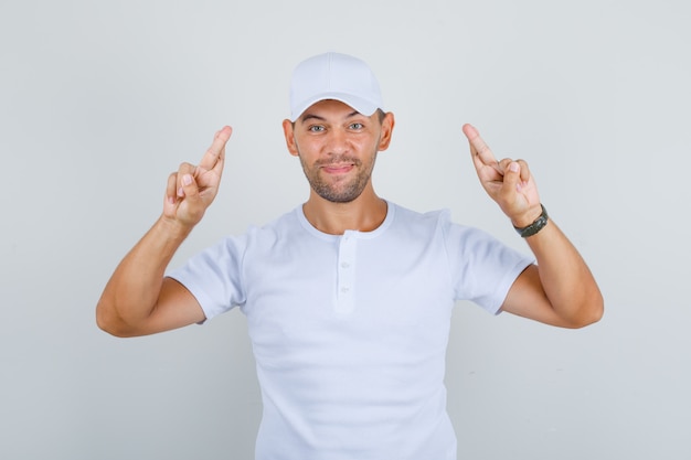 Man gesturing finger crossed and wishing luck in white t-shirt, cap and looking happy, front view.