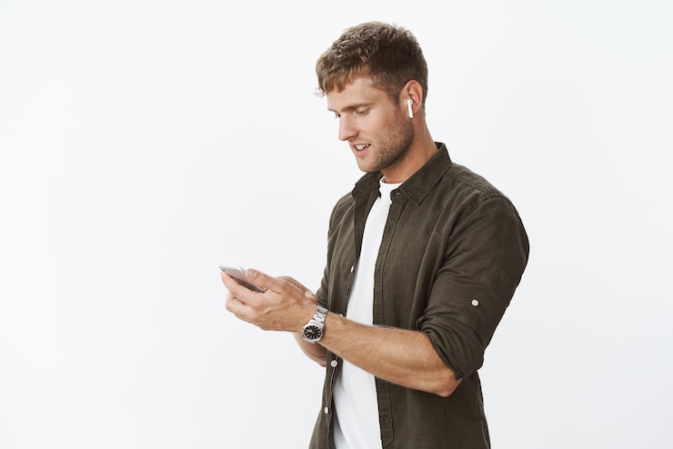 Free Photo man getting outside wearing wireless earphones holding smartphone looking at cellphone screen, listening music, calling friend while on way meeting.