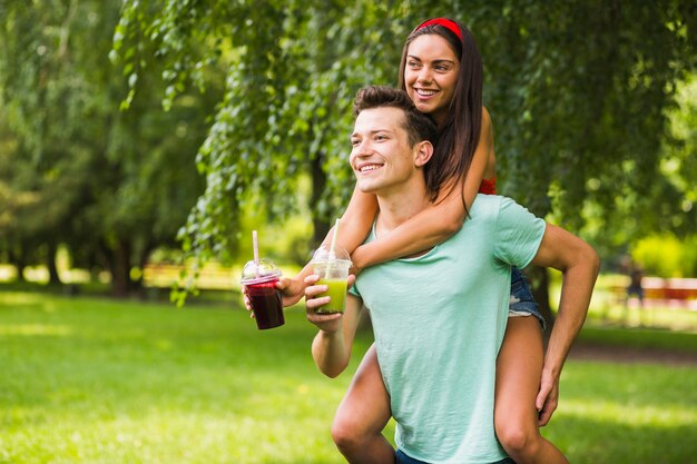Man giving her girlfriend piggyback ride holding smoothies in the park