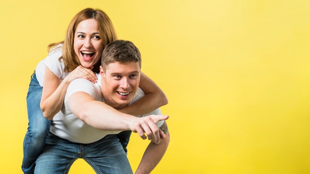 Man giving her laughing girlfriend piggyback ride pointing at camera against yellow background