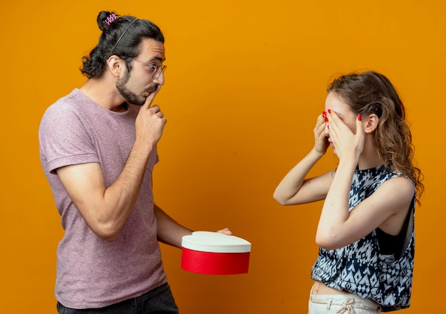 man giving making surprise giving present box to his girlfried while she closing her eyes, young beautiful couple man and women over orange wall